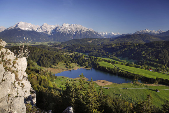 Blick vom Berg auf einen Bergsee und die dahinter liegende Berglandschaft.